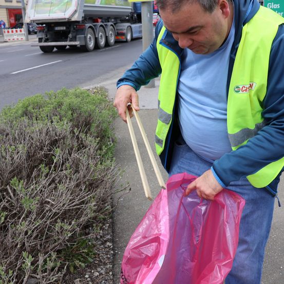 Ein Mann in einer Warnweste sammelt Müll vom Straßenrand auf. Er hält einen Plastikbeutel und einen Stock. Hinter ihm steht ein Lkw auf der Straße.