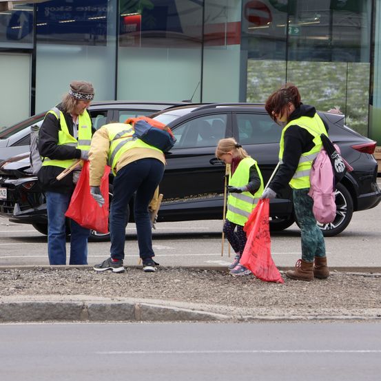 Vier Personen in gelben Warnwesten sammeln Müll am Straßenrand auf. Eine Person beugt sich nach unten, eine andere hält eine rote Tasche, und zwei stehen. Ein schwarzes Auto ist dahinter geparkt.