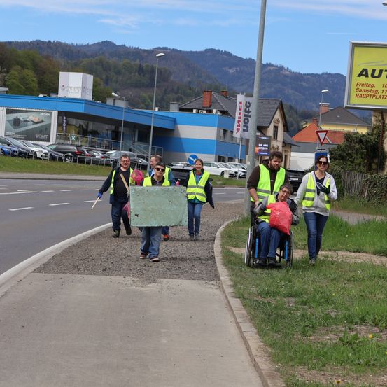 Freiwillige in Warnwesten reinigen eine Straße. Eine Person ist in einem Rollstuhl und trägt einen roten Beutel. Sie befinden sich am Straßenrand mit einem Grasbereich und geparkten Autos. Berge und ein Gebäude sind im Hintergrund.