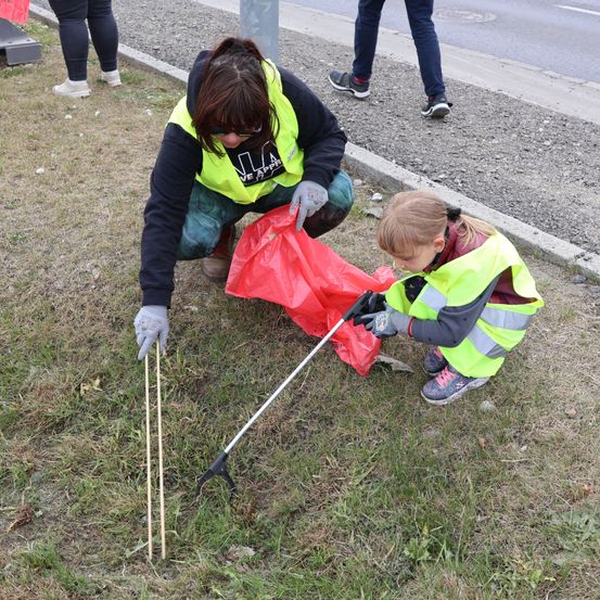Eine Frau und ein Kind sammeln Müll am Straßenrand auf. Beide tragen Warnwesten und Handschuhe. Die Frau benutzt einen Müllgreifer, um den Abfall einzusammeln.