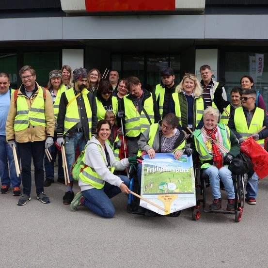 Eine Gruppe von Menschen in Warnwesten und mit Besen posiert für ein Foto vor einem Gebäude, wobei eine Frau ein Plakat hält.