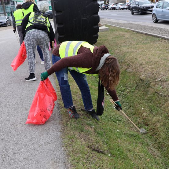 Eine Frau in einer Warnweste sammelt Müll am Straßenrand, hält einen Plastikbeutel und einen Pflücker. Zwei weitere Personen stehen hinter ihr.