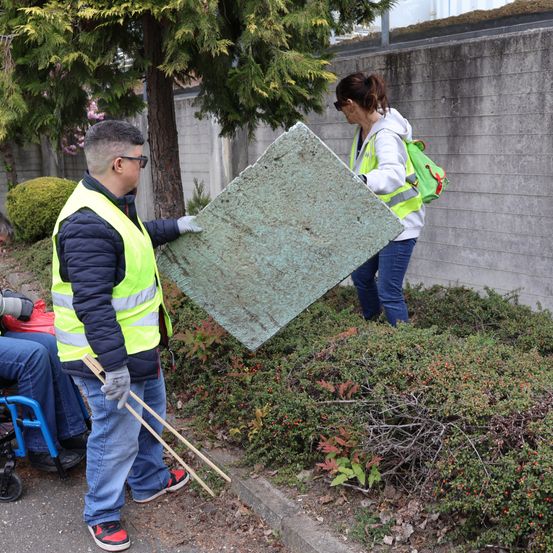 Ein Mann und eine Frau in Sicherheitswesten tragen ein großes Stück grünes Material. Sie stehen in einem Garten mit Pflanzen und einer Betonmauer. Eine Person im Rollstuhl ist in der Nähe.