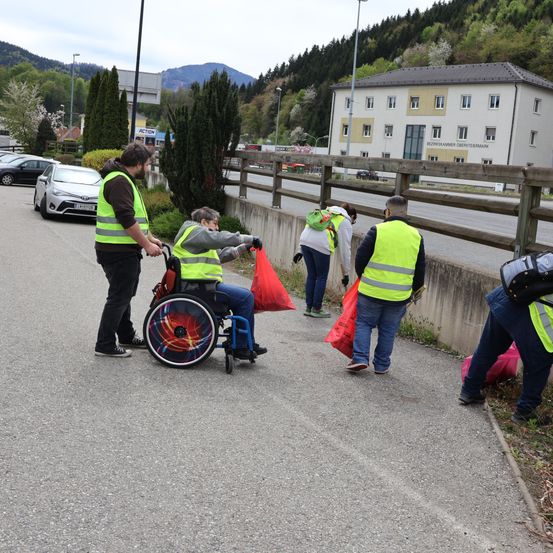 Eine Gruppe von Freiwilligen, darunter eine Person im Rollstuhl, sammelt Müll am Straßenrand. Sie tragen Warnwesten und tragen Säcke. Ein Gebäude und geparkte Autos sind im Hintergrund zu sehen.