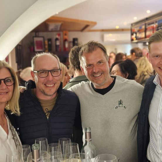 Vier Personen lächeln und stehen vor einem Tisch mit Weingläsern und Flaschen. Ein Mann trägt ein Logo-Shirt, während die anderen lässig gekleidet sind. Dahinter stehen verschwommene Menschen bei einer Zusammenkunft.