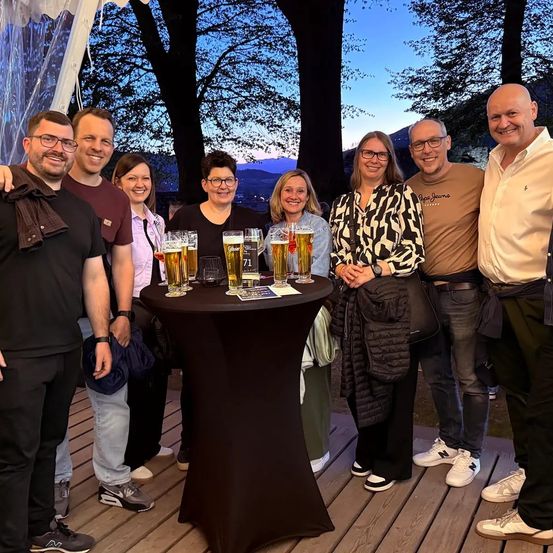 Eine Gruppe von Menschen steht um einen Tisch mit Biergläsern und lächelt für ein Foto. Sie befinden sich auf einer Holzterrasse mit Bäumen und Bergen im Hintergrund.
