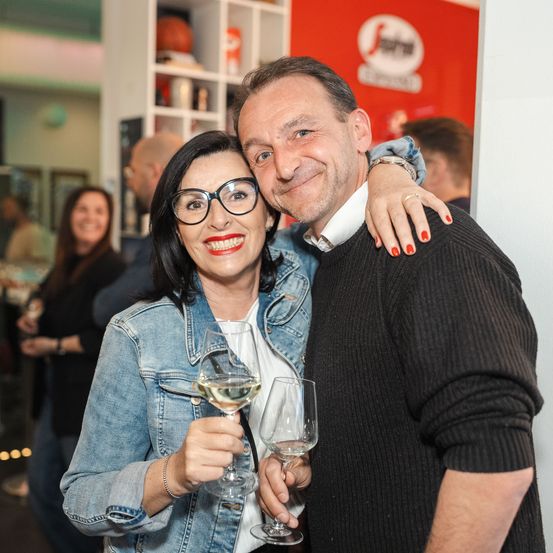 Ein Mann und eine Frau halten Weinglas und lächeln, mit einer anderen Frau und Leute im Hintergrund.