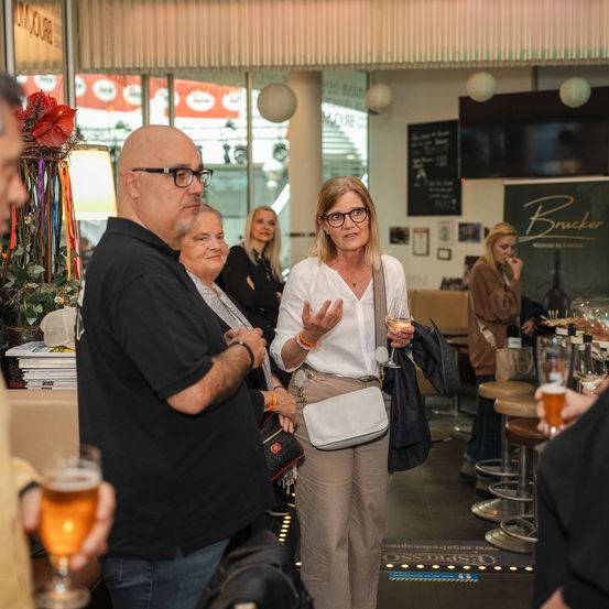 Eine Gruppe von Erwachsenen steht in einer Kneipe, unterhält sich und trinkt Bier. Eine Frau in der Mitte hält ein Glas und spricht. Ein Menübrett mit dem Namen Brucker ist an der Wand.