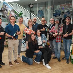 Eine Gruppe von Menschen posiert für ein Foto in einem Raum. Einige halten Bierflaschen in der Hand. Der Mann in der Mitte lächelt und gibt ein Daumen hoch.
