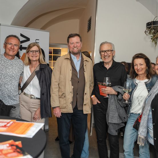 Fünf Erwachsene stehen lächelnd in einem Raum zusammen. Zwei Männer und drei Frauen, alle mit Brille. Ein Mann hält ein Weinglas. Ein Tisch mit Broschüren ist im Vordergrund.