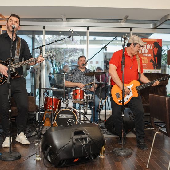 Eine Band spielt auf der Bühne mit Schlagzeug und E-Gitarren. Der Leadsänger spielt die Gitarre, während der Schlagzeuger in der Mitte sitzt. Das Publikum sitzt hinter ihnen.