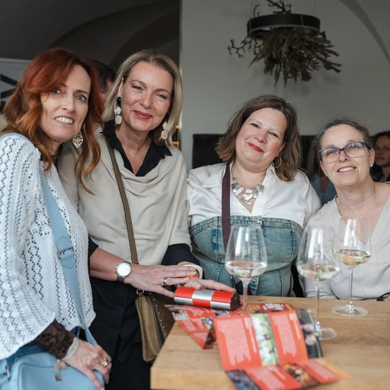 Vier Frauen stehen bei einem Tisch, lächeln und posieren für ein Foto. Sie halten Weingläser und ein Büchlein. Eine dekorative Pflanze hängt an der Decke im Hintergrund.