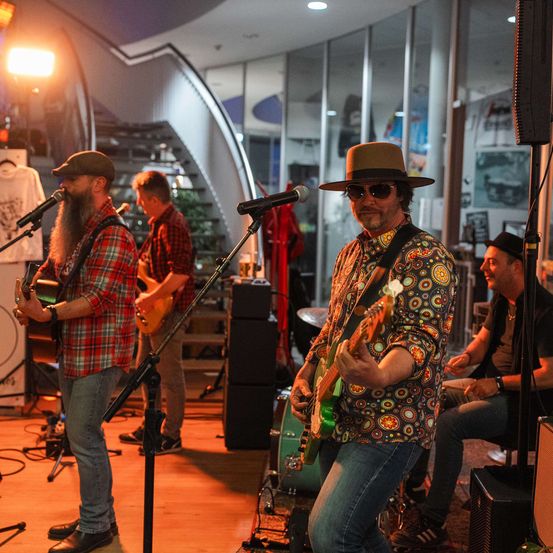 Eine Band spielt auf der Bühne mit Gitarren und einem Schlagzeug, mit einem Mikrofon und Lautsprechern vor ihnen. Dahinter befindet sich eine Treppe und einige Kunstwerke an der Wand.