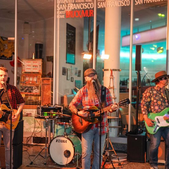 Drei Männer spielen auf einer Bühne mit Musikinstrumenten. Die zentrale Figur spielt Gitarre, während die anderen Gitarre und Schlagzeug spielen. Die Szene spielt in einem Raum mit Glaswänden.