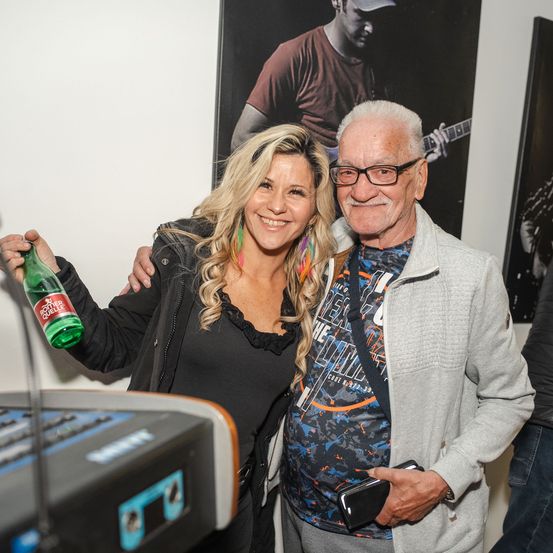 Eine lächelnde Frau, die eine grüne Flasche hält, steht neben einem älteren Mann und beide posieren für ein Foto vor einem Gitarrenposter.