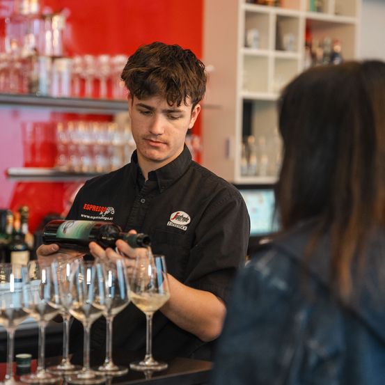 Ein Barkeeper schenkt Wein in Weingläser an einer Bar. Im Hintergrund befinden sich Regale mit verschiedenen Flaschen.