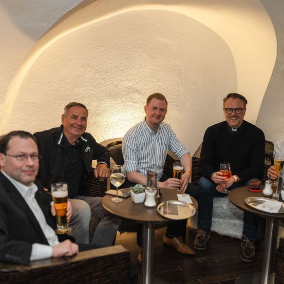 Fünf Männer sitzen in einem schwach beleuchteten Raum und halten Gläser mit Bier und Wein. Sie lächeln und scheinen eine Zusammenkunft zu genießen. Der Raum hat gewölbte Decken und ist mit Tischen und Stühlen eingerichtet.