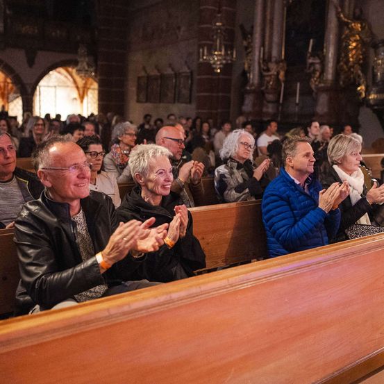 Eine Gruppe von Menschen sitzt auf hölzernen Kirchenbänken und klatscht. Einige tragen Brillen, Armbänder und Jacken. Die Kirche hat eine hohe Decke mit Kronleuchtern.