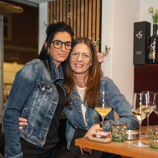 Zwei Frauen sitzen an einem Restauranttisch und lächeln. Eine trägt eine Jeansjacke, die andere eine Brille. Hinter ihnen zeigt eine Wand eine Weinflasche und ein Blumenarrangement.