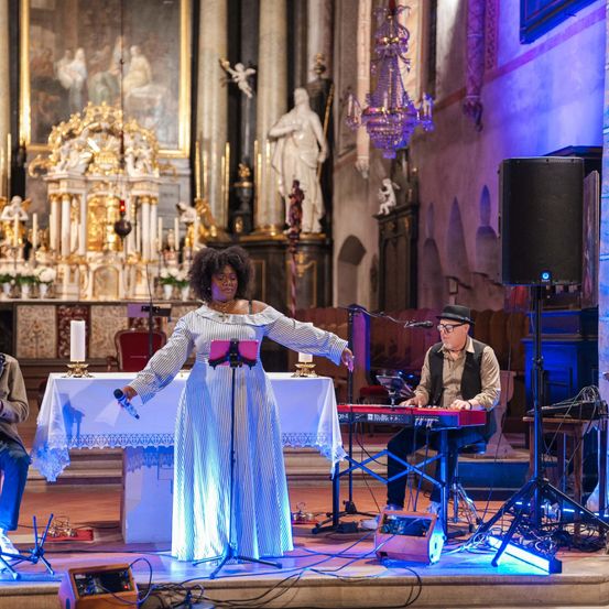 Eine Frau steht mit einem Mikrofon und einem Kleid auf der Bühne in einer Kirche. Ein Mann spielt hinter ihr Klavier. Ein Tisch mit Kerzen und einem Lautsprecher steht auf der Bühne.