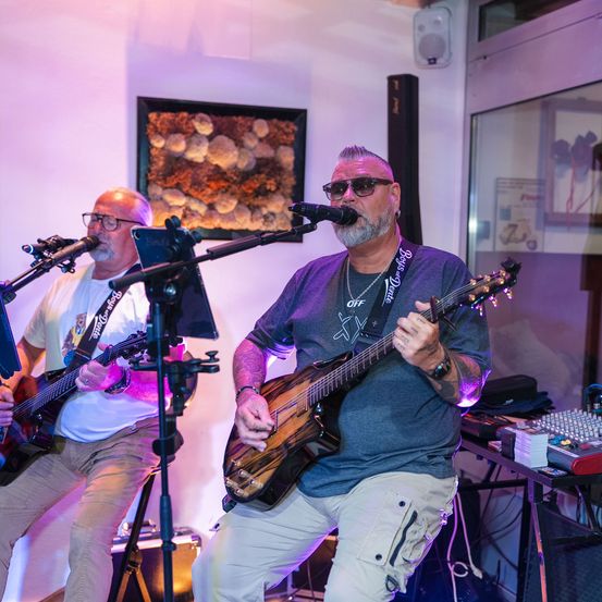 Zwei Männer spielen auf der Bühne, einer spielt Gitarre und singt in ein Mikrofon, der andere spielt Gitarre. Ein Bilderrahmen ziert die Wand hinter ihnen.