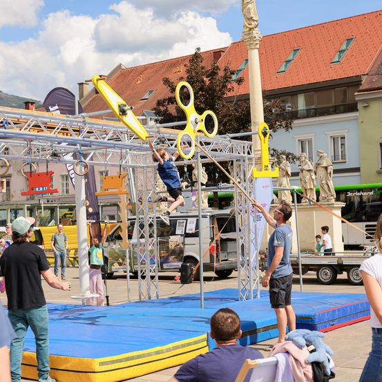 Ein Kind führt eine Kletteraufgabe auf einem Stadtplatz vor, mit Zuschauern, einem Trainer und Skulpturen im Hintergrund.