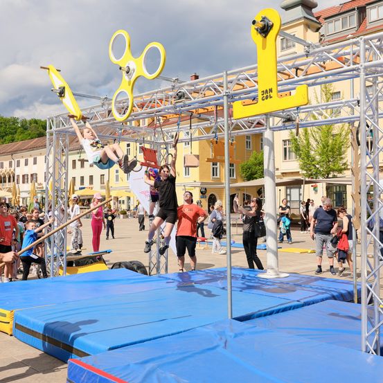 Ein Outdoor-Event, bei dem Menschen auf gelben Strukturen klettern und schwingen. Zuschauer beobachten und fotografieren. Gebäude und Bäume im Hintergrund.