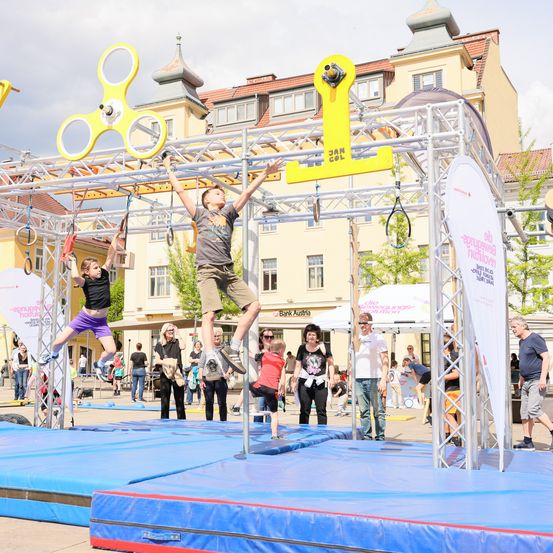 Zwei Kinder schwingen an einer gelben Struktur mit Ringen. Mehrere Menschen beobachten sie. Die Umgebung ist draußen, mit Gebäuden und Bäumen im Hintergrund.