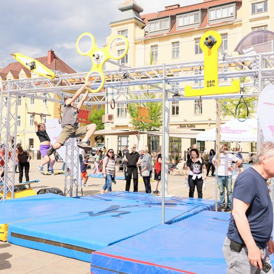 Menschen nehmen an einer körperlichen Herausforderung teil. Zwei Personen hängen an einer gelben Struktur, während andere im Hintergrund stehen. Ein Mann beobachtet genau.
