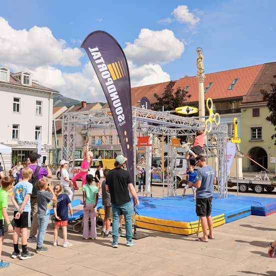 Eine Gruppe von Menschen beobachtet einen Mann, der einen Stunt auf einer Plattform auf dem Platz einer Stadt durchführt, mit einem großen Banner und Gebäuden im Hintergrund.