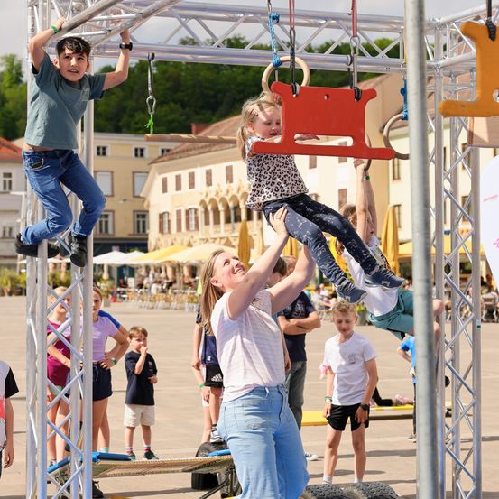Eine Frau stützt ein Mädchen, das an einem Barren hängt, während ein Junge auf einem anderen Barren klettert in einer Outdoor-Spielstruktur. Andere Kinder beobachten und Erwachsene stehen in der Nähe. Gebäude und Grünflächen sind im Hintergrund.