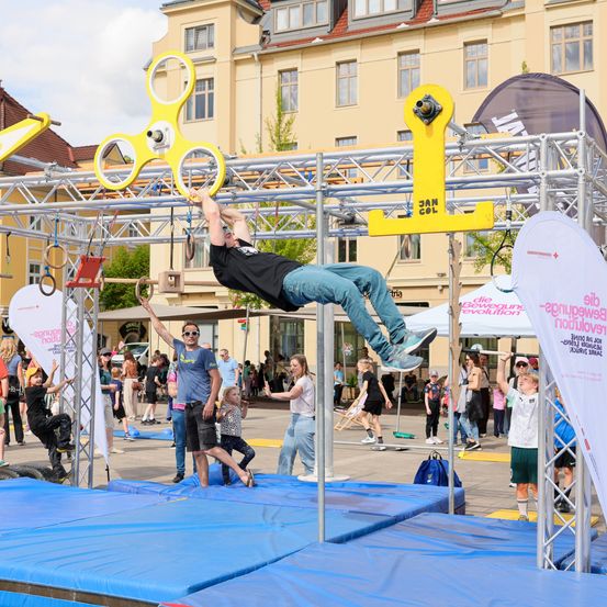 Ein Mann in einem schwarzen Shirt führt einen Stunt auf einer gelben Plattform mit einem gelben Spinner durch, während andere im Hintergrund zuschauen.