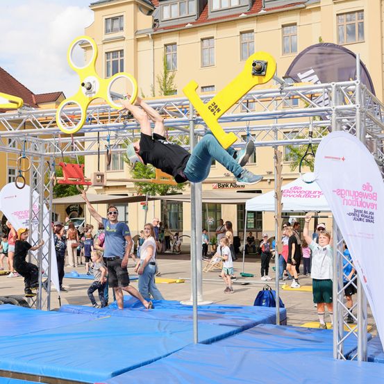 Ein Mann führt Akrobatik auf einer Metallkonstruktion mit gelben Werkzeugen und Ringen bei einer Veranstaltung im Freien vor. Zuschauer beobachten vom Straßenrand, einige stehen auf blauen Matten. Ein Gebäude mit vielen Fenstern ist im Hintergrund.