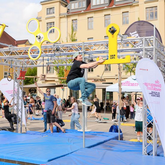 Ein Mann klettert auf einer Hindernislauf-Struktur bei einer Outdoor-Veranstaltung. Er hält sich an einem gelben Balken fest, während andere zusehen. Im Hintergrund sind Gebäude und Menschen zu sehen.