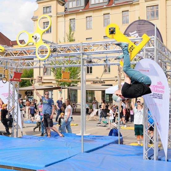 Ein Mann vollführt einen Handstand auf einer gelben Metallkonstruktion mit Ringen und einem Banner. Viele Menschen, einschließlich Kinder, beobachten ihn an einem sonnigen Tag. Die Szene spielt im Freien, mit Gebäuden und Bäumen im Hintergrund.