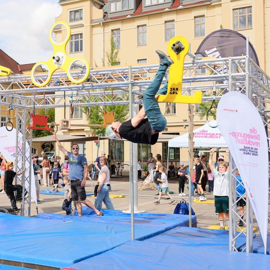 Ein Mann vollführt einen Handstand auf einer Metallkonstruktion bei einer Outdoor-Veranstaltung. Zuschauer beobachten von den Seiten. Die Konstruktion ist gelb und silber, mit verschiedenen Gegenständen, die daran hängen. Im Hintergrund befindet sich ein gelbes Gebäude.