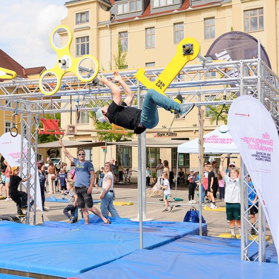 Ein Athlet führt einen Handstand auf einer Metallkonstruktion bei einer Outdoor-Veranstaltung aus. Zuschauer beobachten hinter Sicherheitsmatten. Banner befinden sich an den Seiten. Gebäude und Bäume sind im Hintergrund.