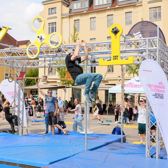 Ein Mann klettert auf einer gelben Stange in einem Außenbereich mit blauen Matten. Mehrere Menschen stehen um ihn herum und beobachten. Es sind auch mehrere Zelte aufgebaut und eine große gelbe Struktur.