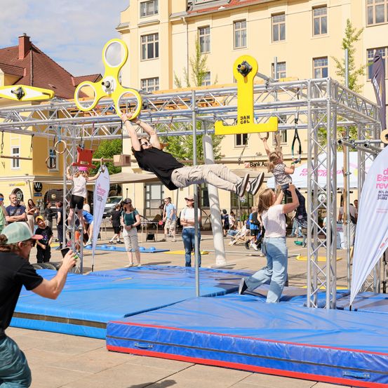 Mehrere Personen beschäftigen sich auf einem Outdoor-Spielplatz mit körperlichen Aktivitäten. Ein Mann hängt an einer Stange, während eine Frau ein Kind hält. Eine andere Frau klettert eine vertikale Leiter. Der Spielplatz ist von Zuschauern umgeben.