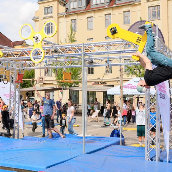 Eine Outdoor-Veranstaltung mit verschiedenen Zirkusnummern. Ein Mann ist kopfüber auf einem Trapez, während andere zusehen. Farbenfrohe Geräte und Banner sind vorhanden.