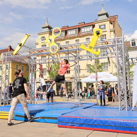 Ein junger Mensch nimmt an einem Hindernisparcours mit einer gelben Struktur und blauen Matten teil. Mehrere Personen beobachten die Veranstaltung im Freien.