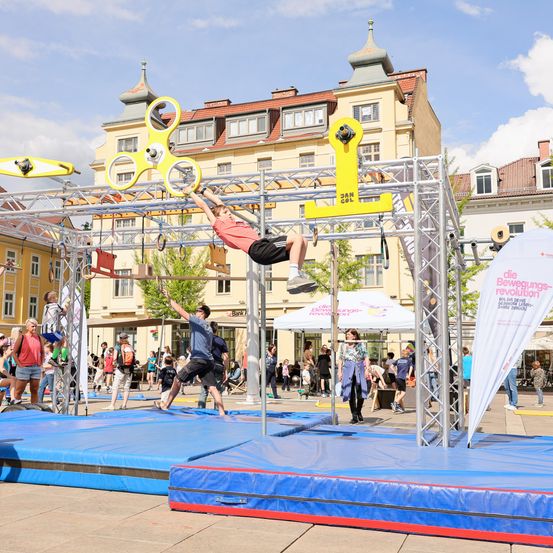 Ein Outdoor-Hindernisparcours mit Teilnehmern, die verschiedene Aktivitäten ausführen, darunter das Hängen an Ringen und das Klettern einer vertikalen Leiter, auf einem Stadtplatz mit Zuschauern.