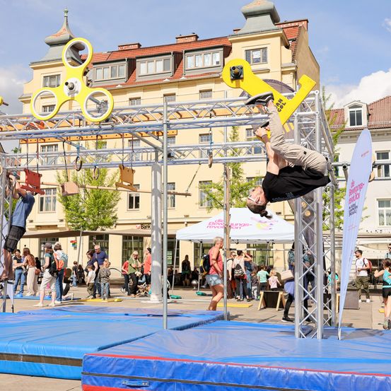 Akrobatik-Vorstellung in einer urbanen Umgebung. Ein Mann hängt kopfüber an einer gelben Konstruktion, während andere zuschauen.