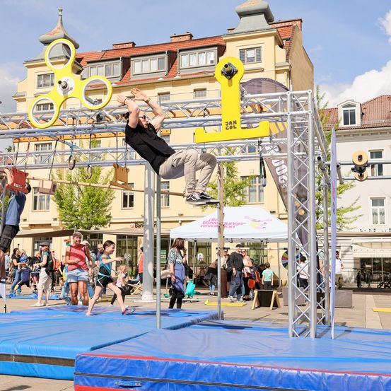 Ein Mann in einem schwarzen T-Shirt und khakifarbenen Hosen führt einen Handstand auf einer Plattform aus. Im Hintergrund gehen Leute herum, und ein Zelt ist aufgestellt. Gebäude mit roten Dächern sind sichtbar.