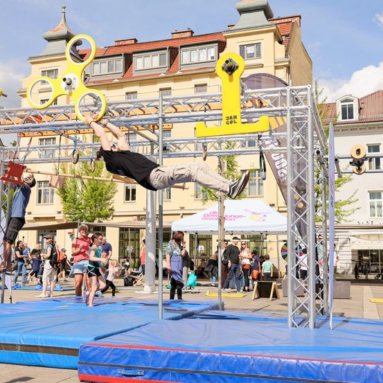 Ein Mann führt einen akrobatischen Stunt auf einem Hindernisparcours im öffentlichen Außenbereich durch. Mehrere Zuschauer beobachten.