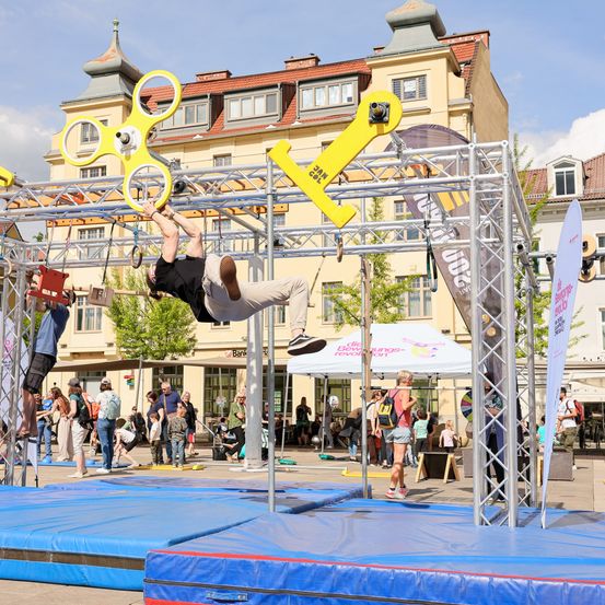 Ein Mann führt einen Stunt auf einem Hindernisparcours mit gelber Ausrüstung im öffentlichen Raum durch. Zuschauer beobachten von unten, und in der Nähe gibt es Zelte und Banner.