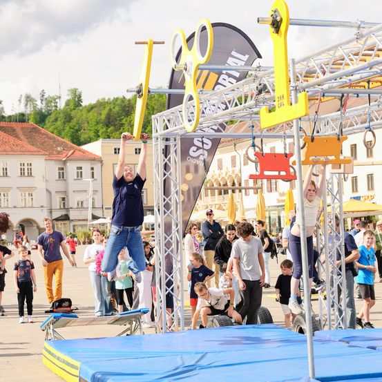 Menschen nehmen an einer Sportveranstaltung im Freien teil, wobei einer an einer Stange hängt und ein anderer auf einer Plattform steht. Im Hintergrund befinden sich Gebäude und Bäume.