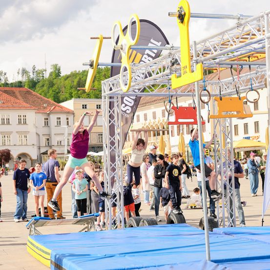 Zwei Kinder klettern auf einem Hindernisparcours, den Zuschauer im Stadtplatz beobachten. Sie greifen nach Ringen.