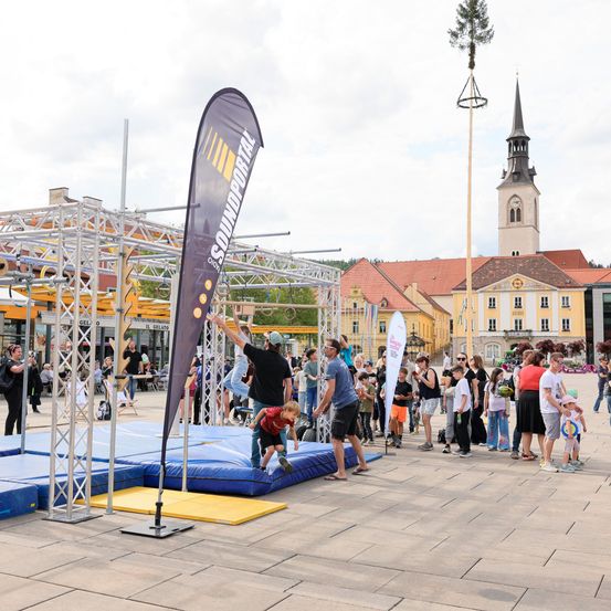 Eine Gruppe von Menschen beobachtet einen Mann und ein Kind, die auf einer Plattform in einem Außenbereich springen. Ein großer Banner steht in der Nähe. Ein Gebäude mit einem Turm und einem Glockenturm ist im Hintergrund.