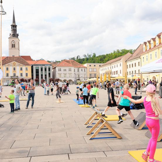 Auf einem offenen Platz nehmen Kinder an einer verspielten Aktivität teil und springen über Holzhürdeln. Im Hintergrund sind Gebäude mit roten Dächern zu sehen, eines davon hat einen Turm.